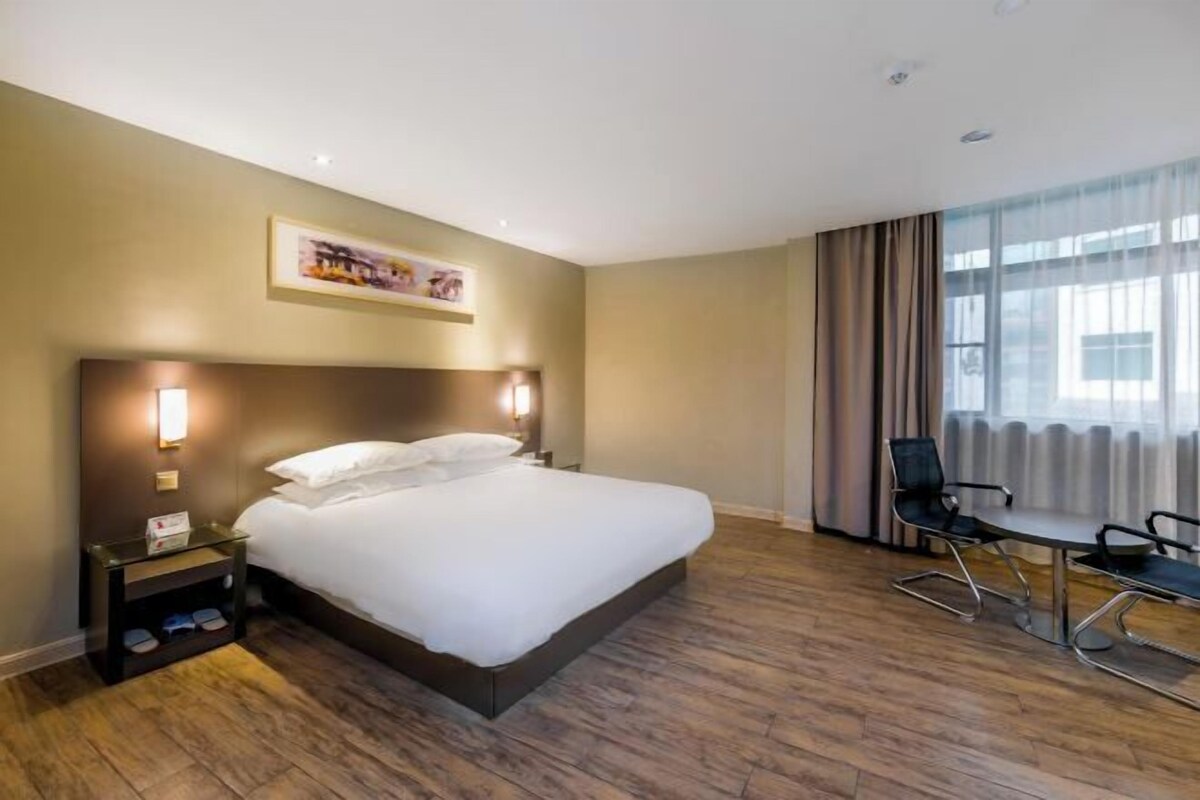 A hotel room with a large bed, bedside tables, a desk with chairs, and a window with curtains.