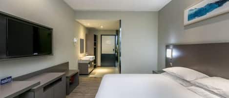 A modern hotel room with a large bed, a flat-screen TV mounted on the wall, a small desk, and a bathroom visible through an open door.