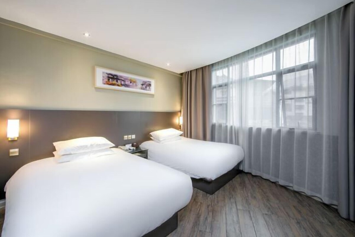 A hotel room with two beds, a large window with sheer curtains, a wall-mounted light, and a framed picture.