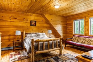 Un dormitorio de cabaña de troncos con una cama de madera, dos lámparas de noche, un sofá y una ventana con vista a árboles.
