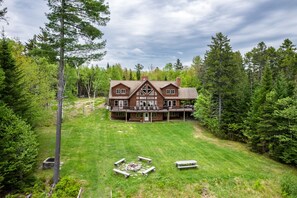 A large, two-story house with a wrap-around porch, surrounded by a well-maintained lawn and dense forest.