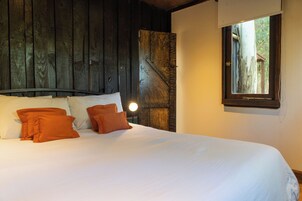 A bed with white linens and orange pillows, a wooden headboard, and a window with a view of trees.