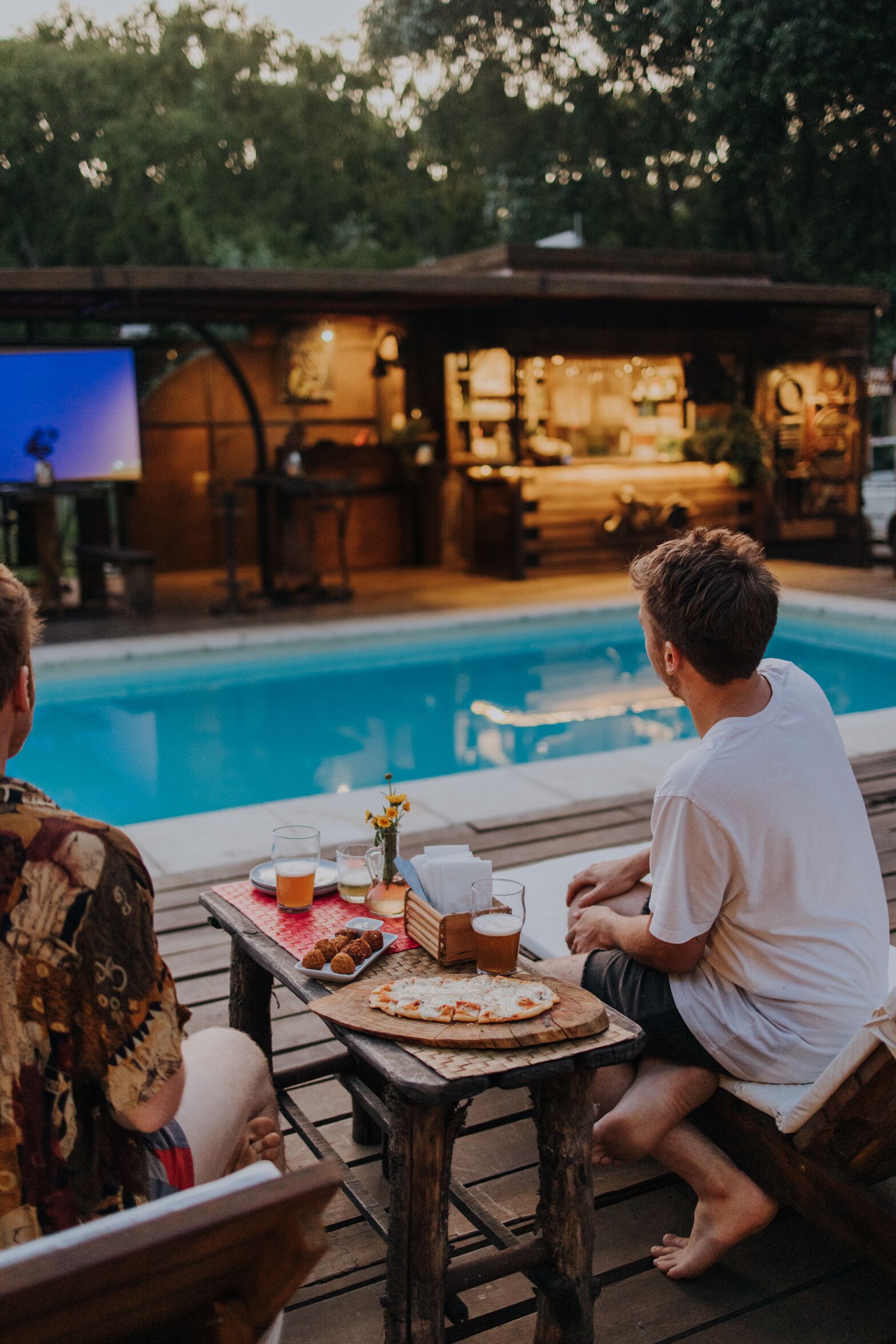 A poolside dining area with a wooden table, a pool, and a bar area in the background.