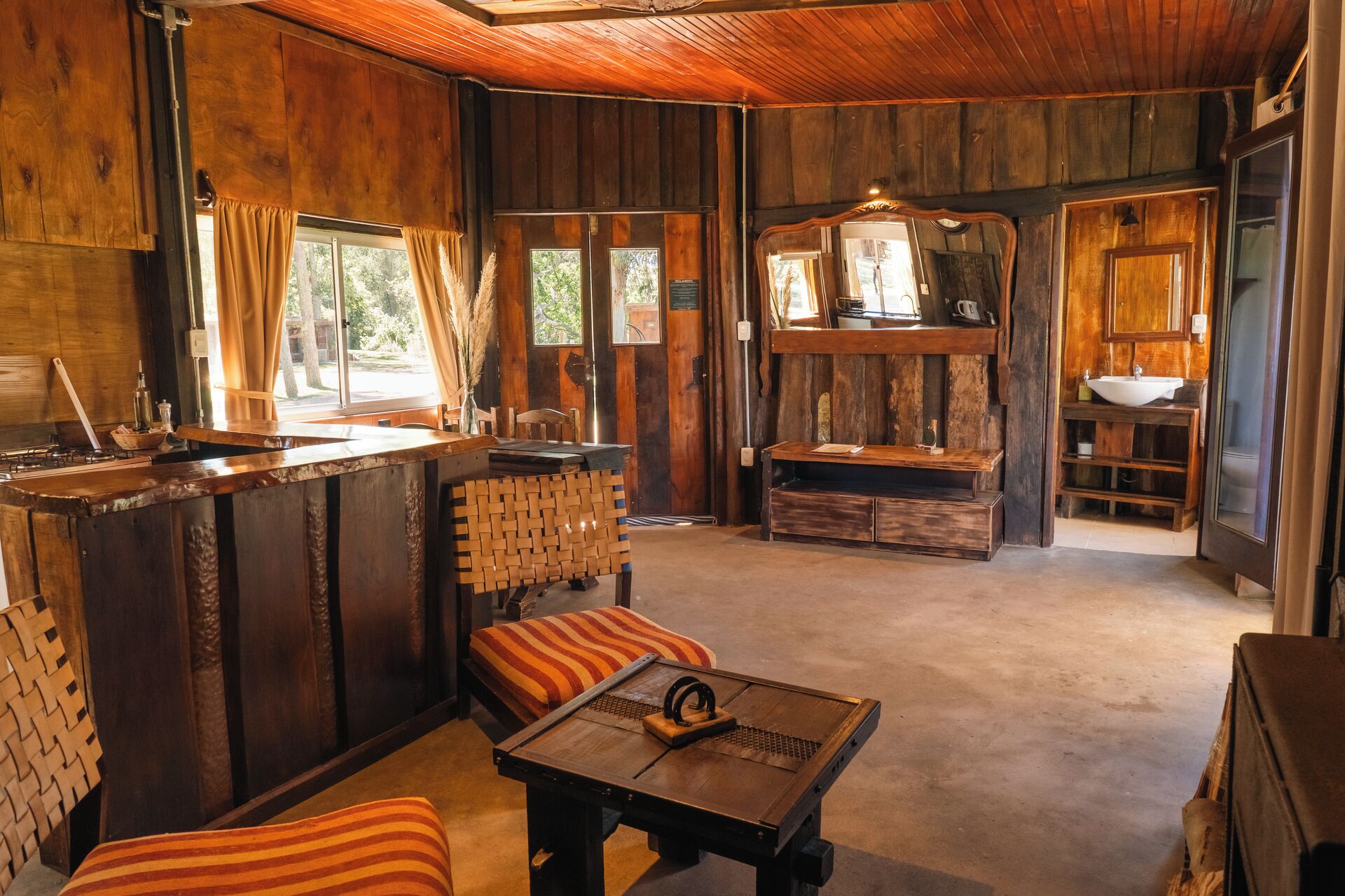 A rustic cabin interior with a wooden bar, seating area, and a bathroom with a large mirror and sink.