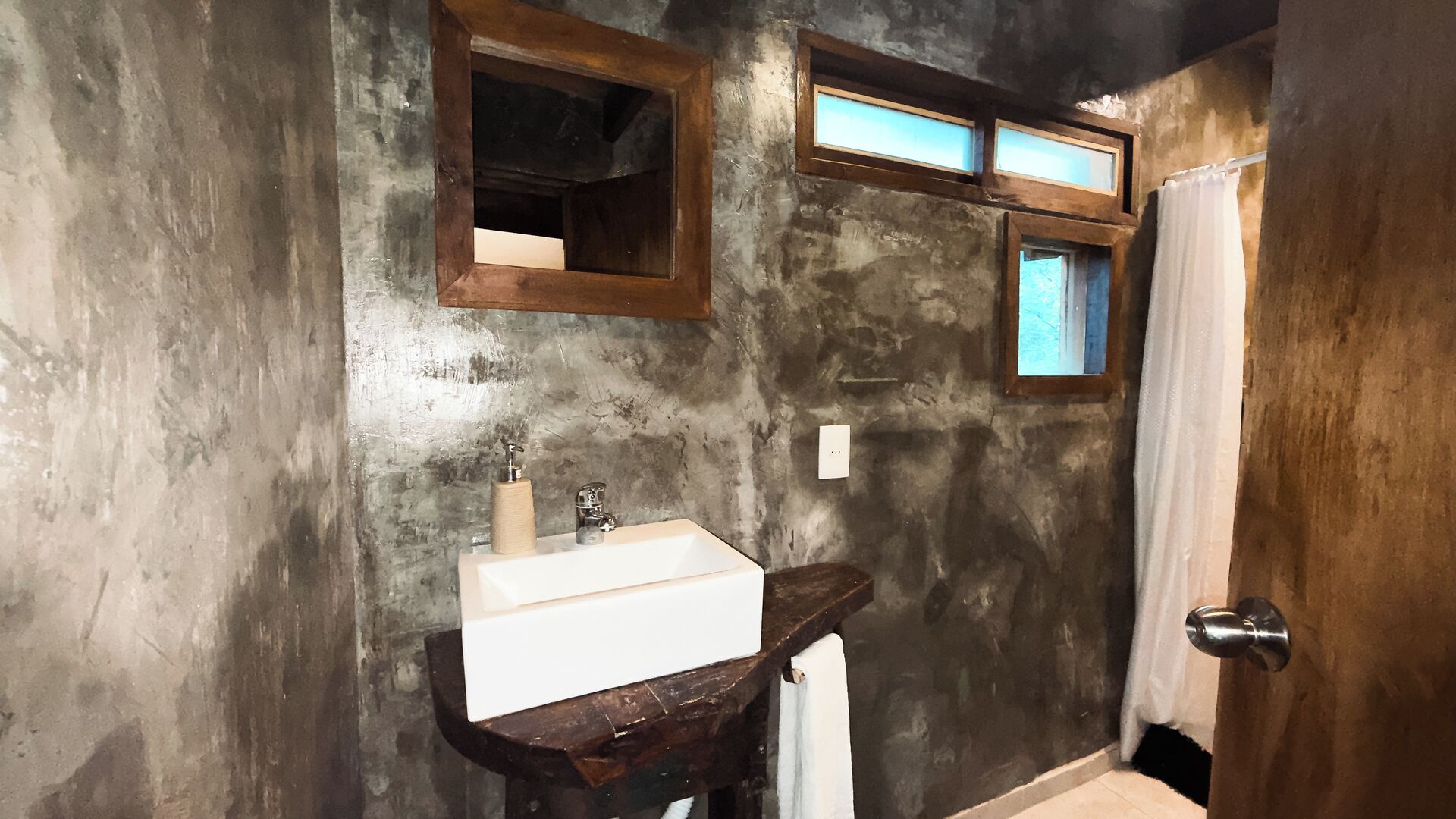 Un baño con pared de concreto rústica, tocador de madera con lavamanos, espejo y ventana con vistas.