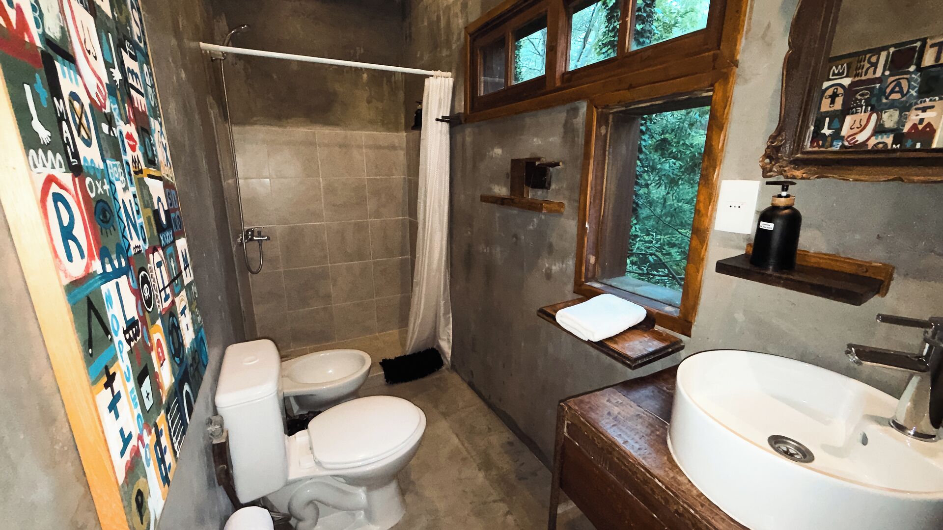 Une salle de bain avec douche, toilettes et lavabo. Il y a une fenêtre donnant sur des arbres.