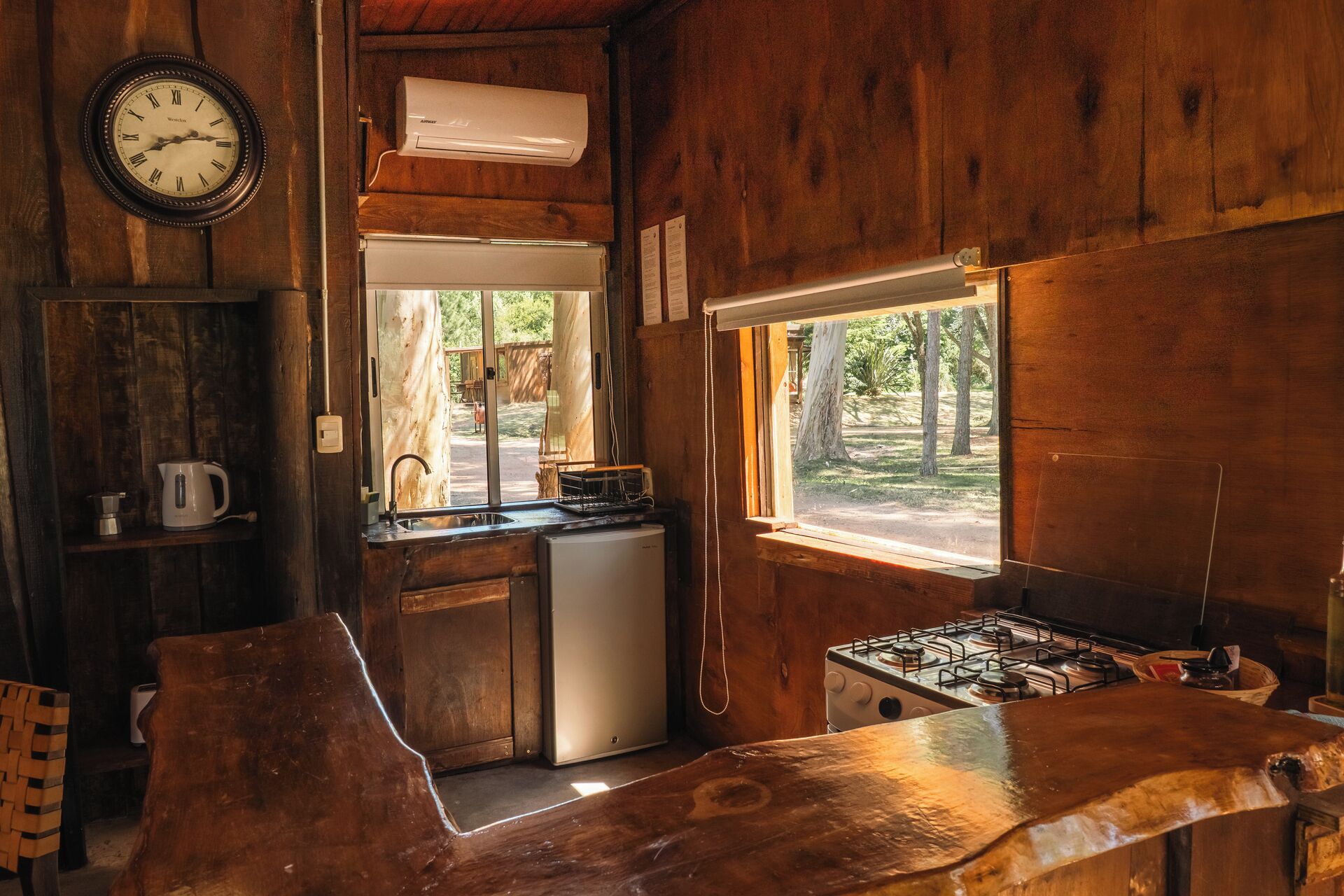 A rustic cabin interior with a wooden interior, a kitchen area with a stove, a refrigerator, and a sink. There is a clock on the wall and a window providing a view of the outdoors.
