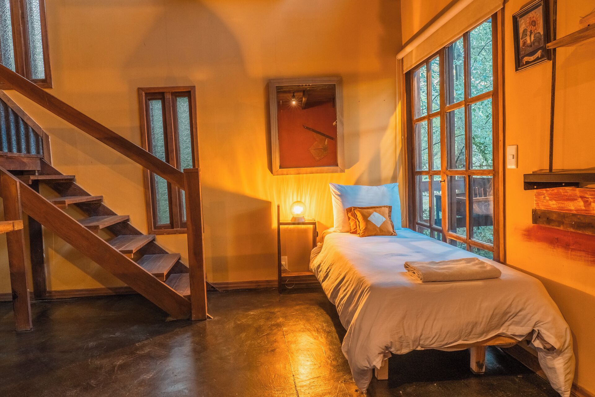 A bedroom with a wooden staircase, a bed with a white duvet, a nightstand with a lamp, and a window with a view of trees.