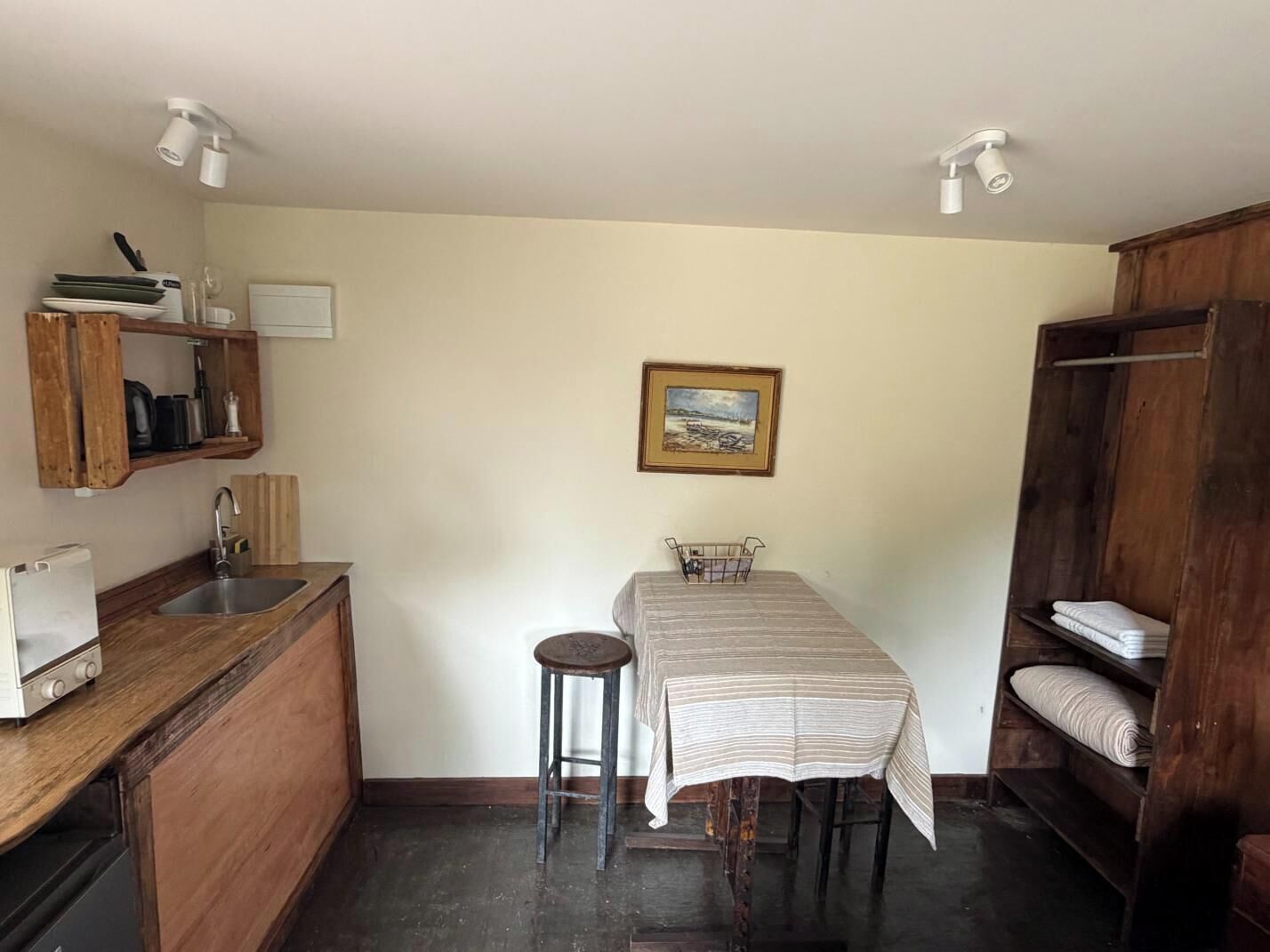 A compact kitchen with a sink, a small dining table, and a stool. There is a painting on the wall and a wooden shelf with towels.