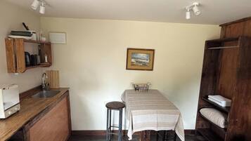 A compact kitchen with a sink, a small dining table, and a stool. There is a painting on the wall and a wooden shelf with towels.