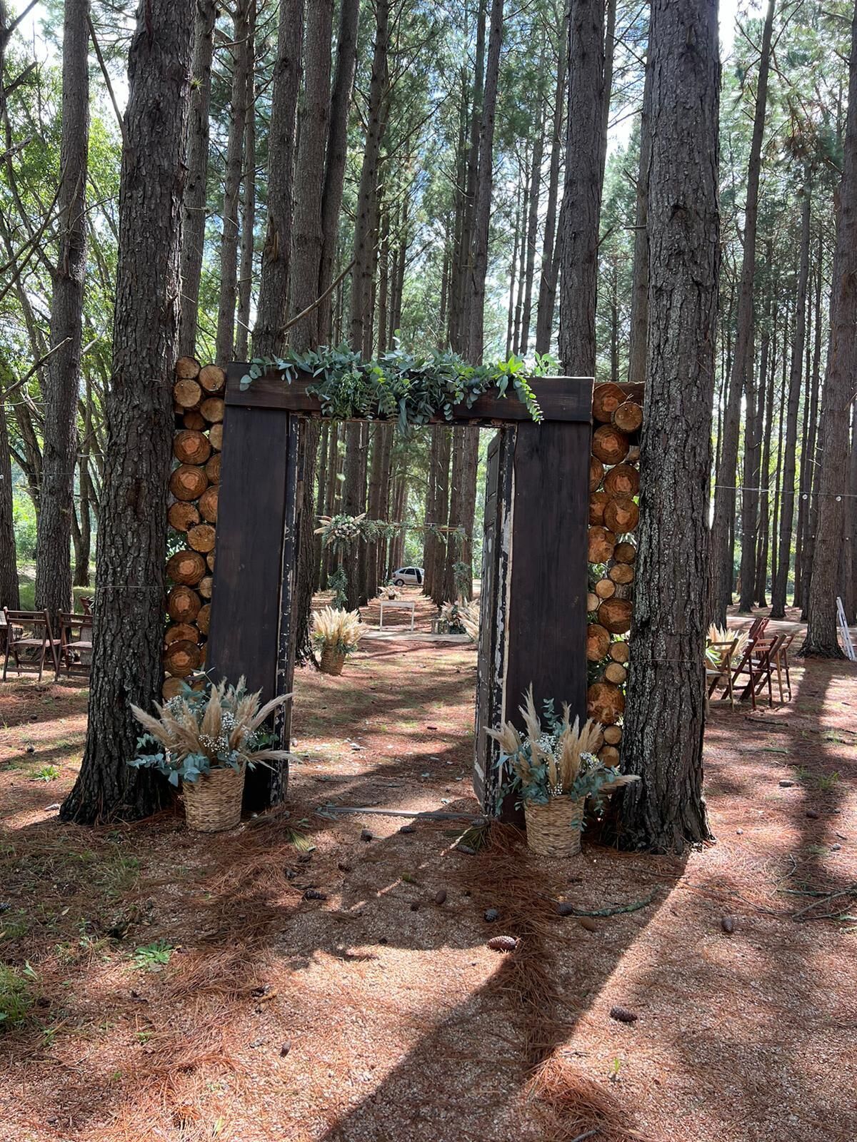 A rustic wooden archway with stacked logs and greenery, surrounded by tall pine trees in a forest setting.