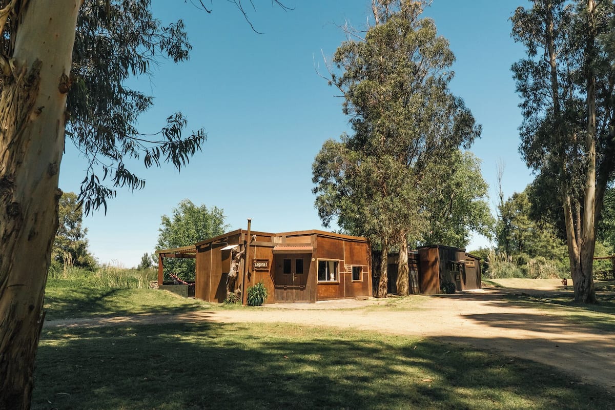 A rustic wooden cabin with a small porch, surrounded by tall trees and a clear sky.