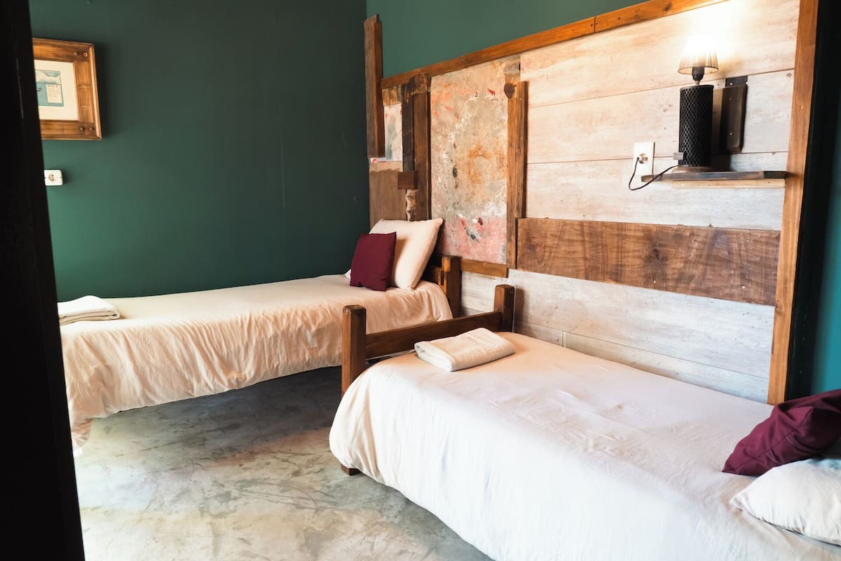 A room with two single beds, each with a wooden headboard and white bedding. There is a wall-mounted shelf with a lamp and a framed picture.