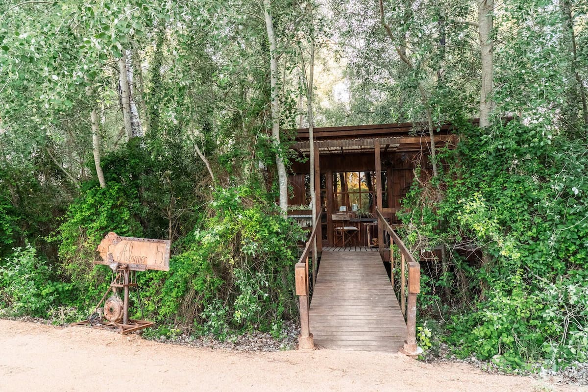 A rustic wooden structure with a wooden staircase leading to a covered seating area surrounded by dense greenery.