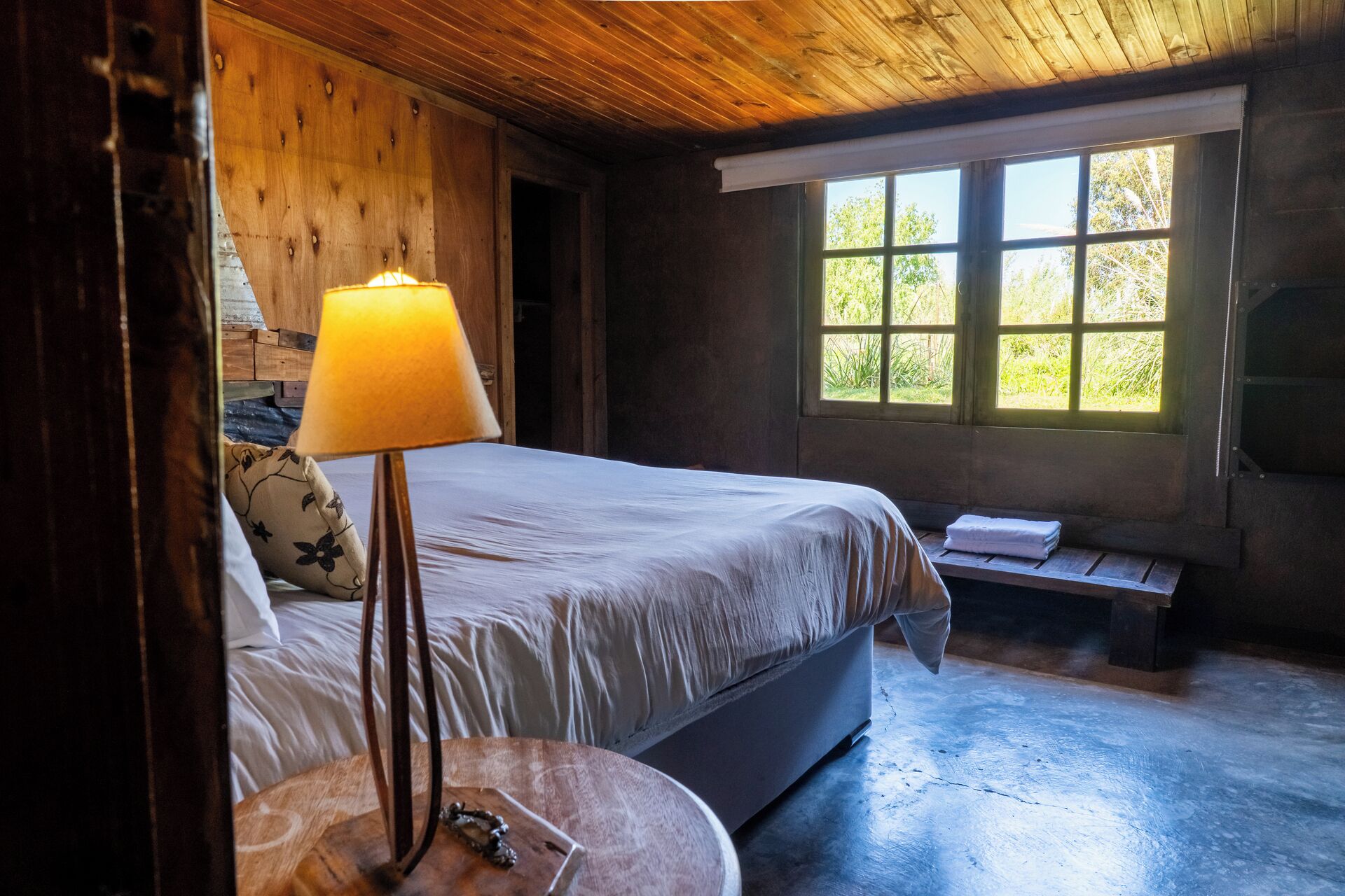 Dormitorio de cabaña de madera con una cama, una lámpara de noche, un banco y una ventana con vista a follaje.