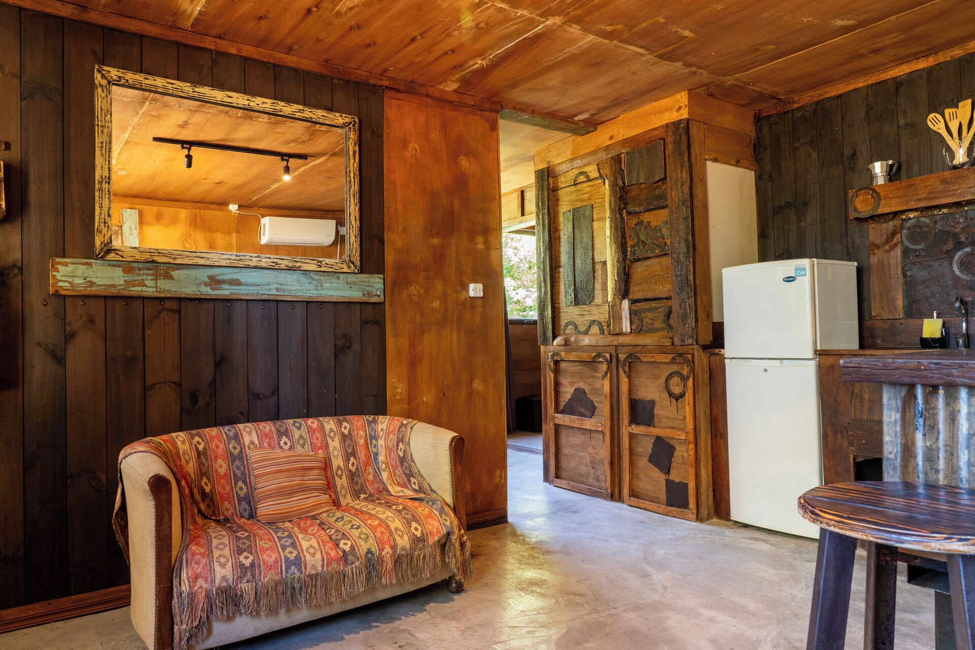 Interior rústico de una cabaña con un sofá, paredes de madera, un espejo, un refrigerador y una mesa de comedor.