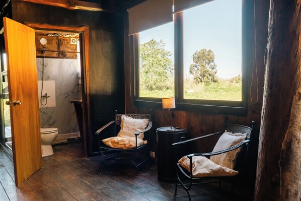 A rustic bathroom with a wooden door, a toilet, and a shower. There are two wicker chairs with cushions and a lamp near a window overlooking a green landscape. - Estancia CH (Maldonado)