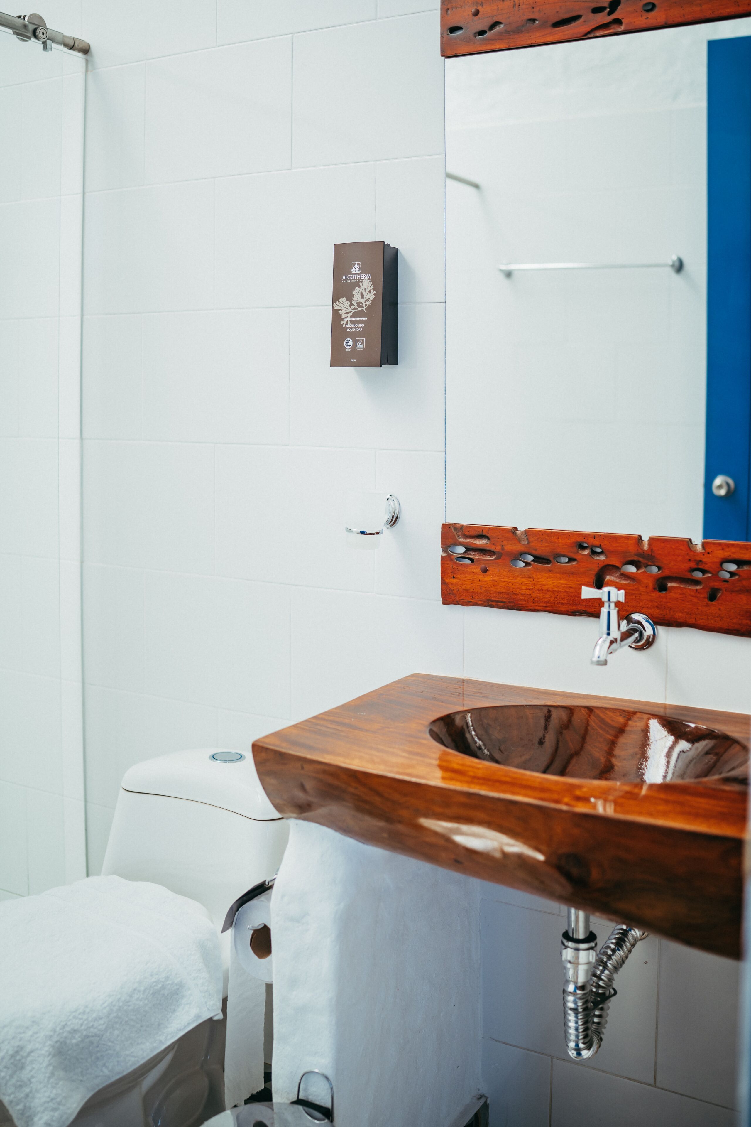 Un baño con un lavamanos de madera, baldosas blancas, una puerta azul y una toalla en el suelo.