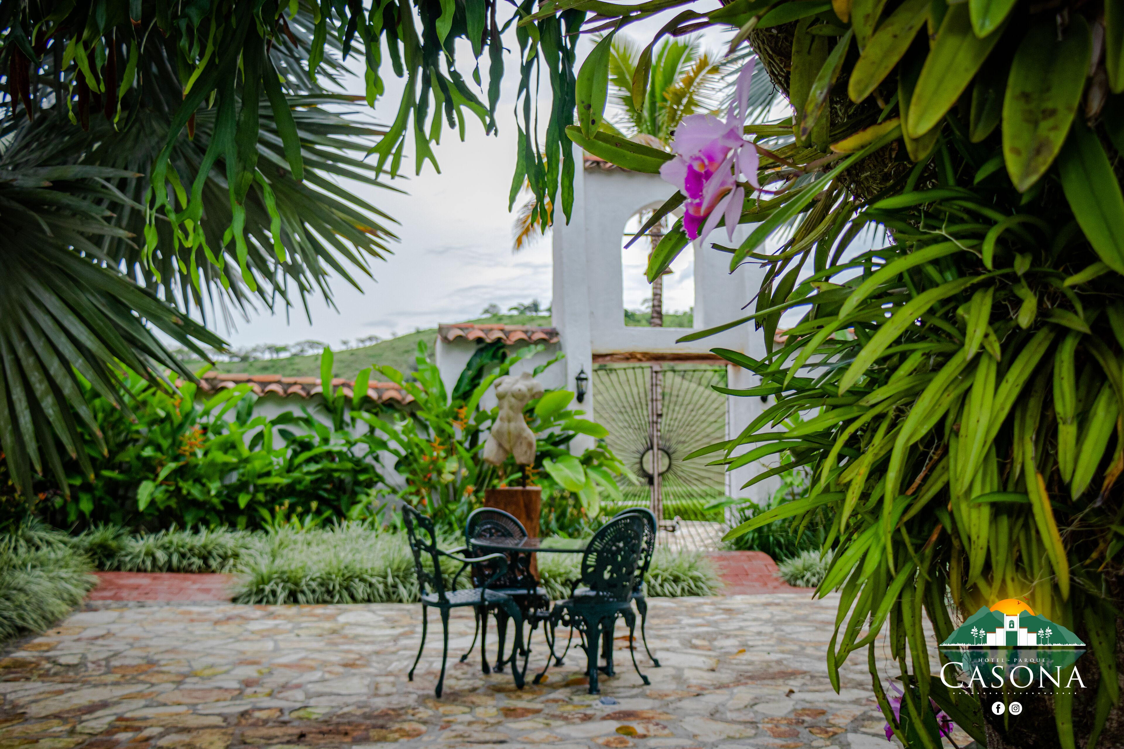 Eine Terrasse mit Steinbelag, zwei Stühle, ein Tisch und üppiges Grün, darunter eine rosa Orchidee.