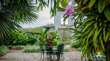 Eine Terrasse mit Steinbelag, zwei Stühle, ein Tisch und üppiges Grün, darunter eine rosa Orchidee.