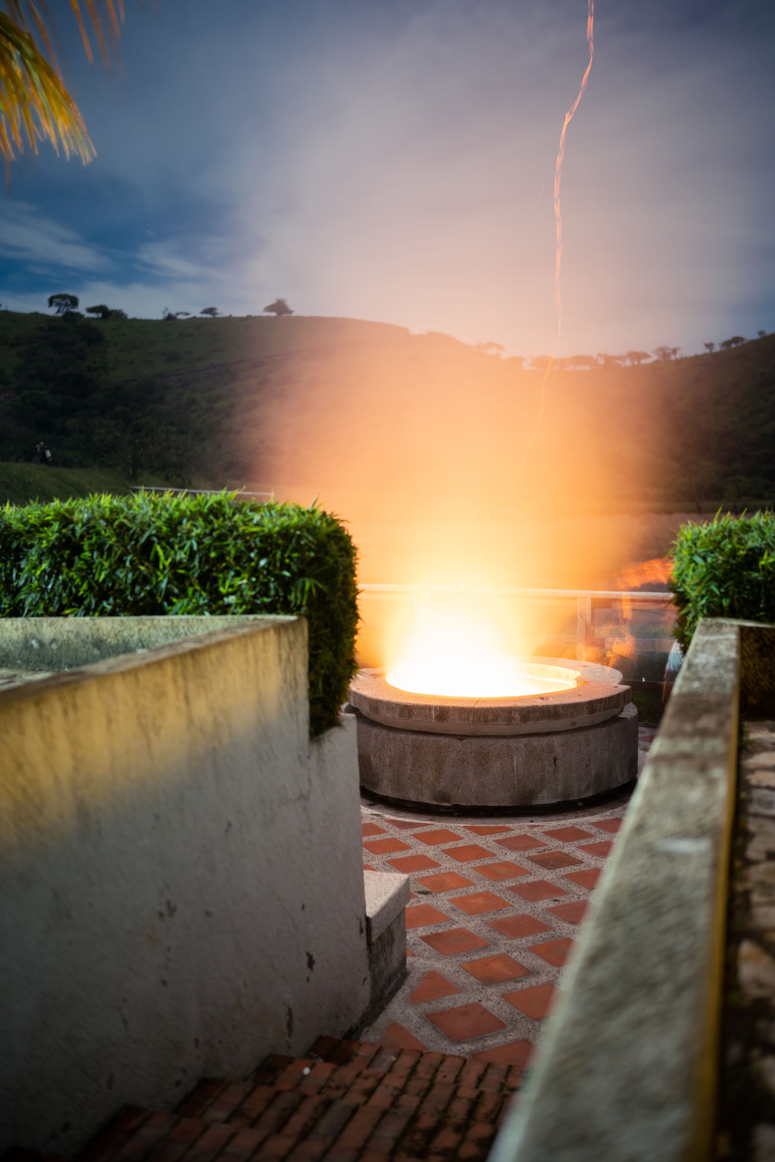 A circular outdoor seating area with a fire feature, surrounded by a low brick wall and greenery.