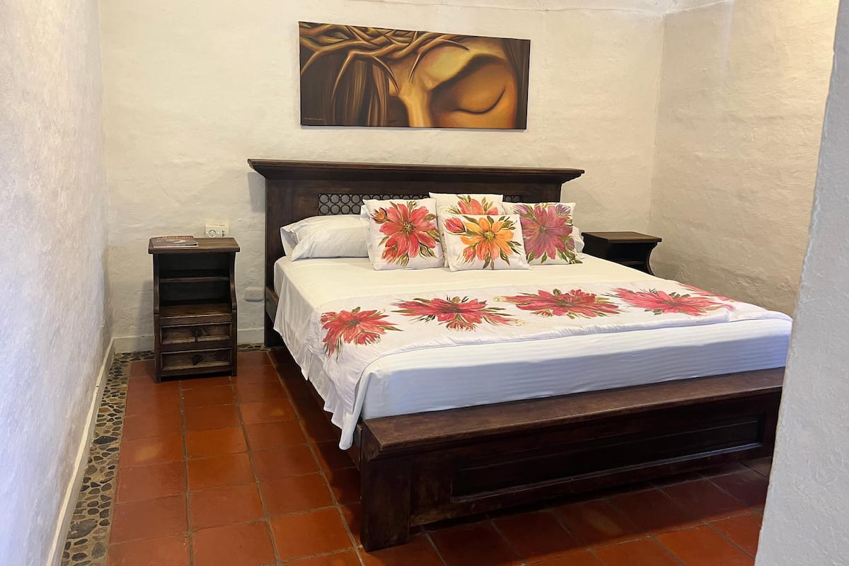 Un dormitorio con cama de madera, ropa de cama blanca, almohadas estampadas, una mesita de noche, un cuadro en la pared y suelo de baldosas.