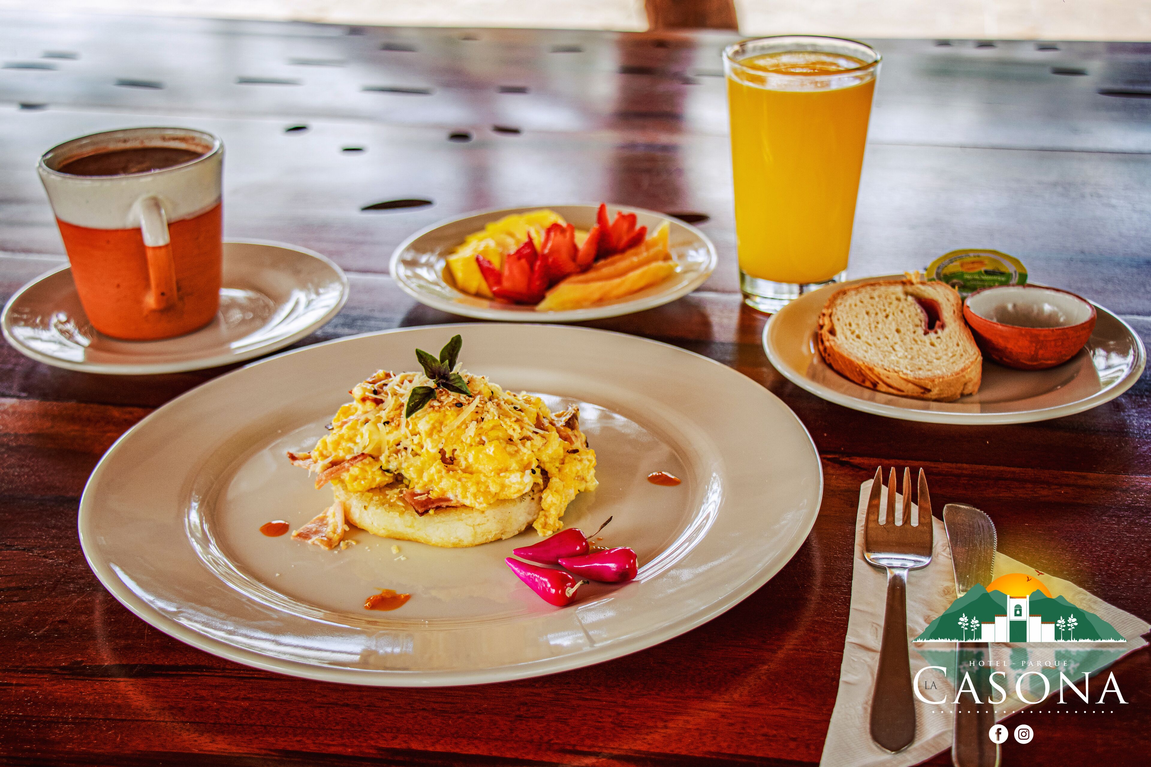 A breakfast setting with a plate of scrambled eggs, toast, and fruit, a glass of orange juice, and a cup of tea on a wooden table.