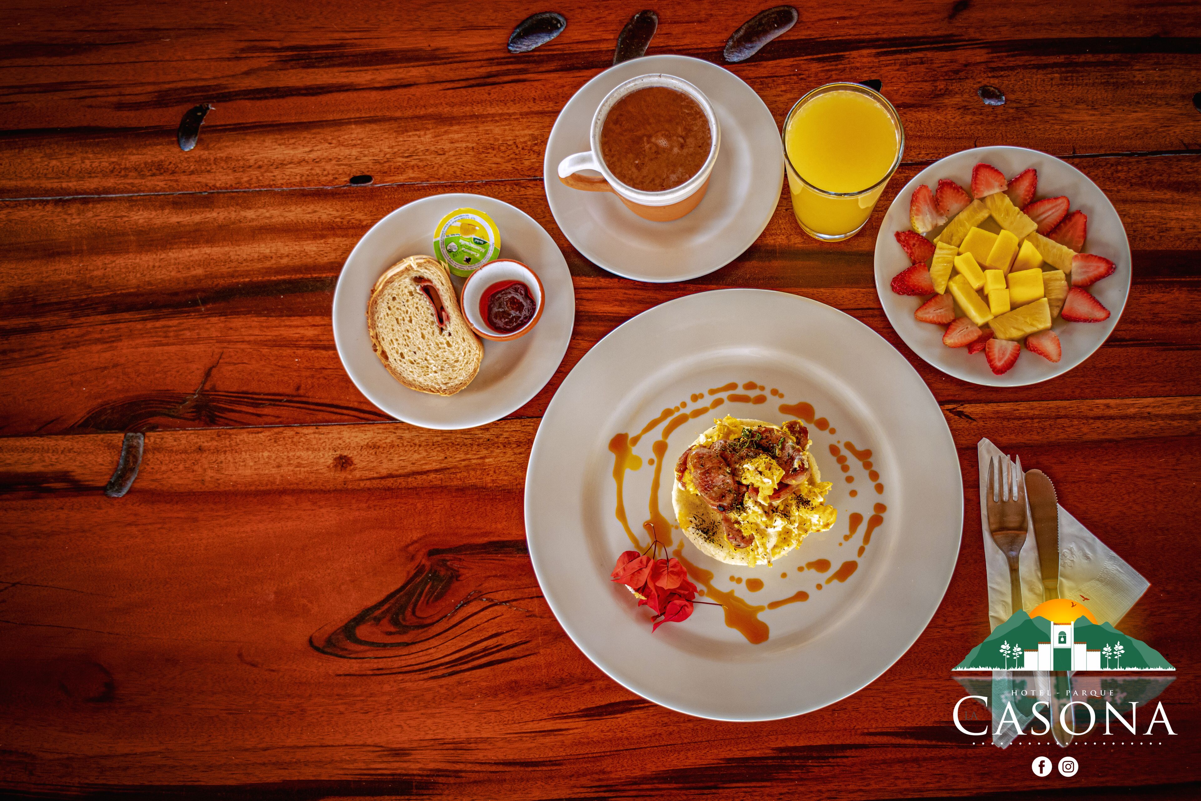 Un desayuno con huevos, tostadas y mermelada, una taza de café, un vaso de jugo de naranja y una fuente de fruta sobre una mesa de madera.