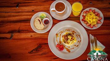 Un desayuno con huevos, tostadas y mermelada, una taza de café, un vaso de jugo de naranja y una fuente de fruta sobre una mesa de madera.