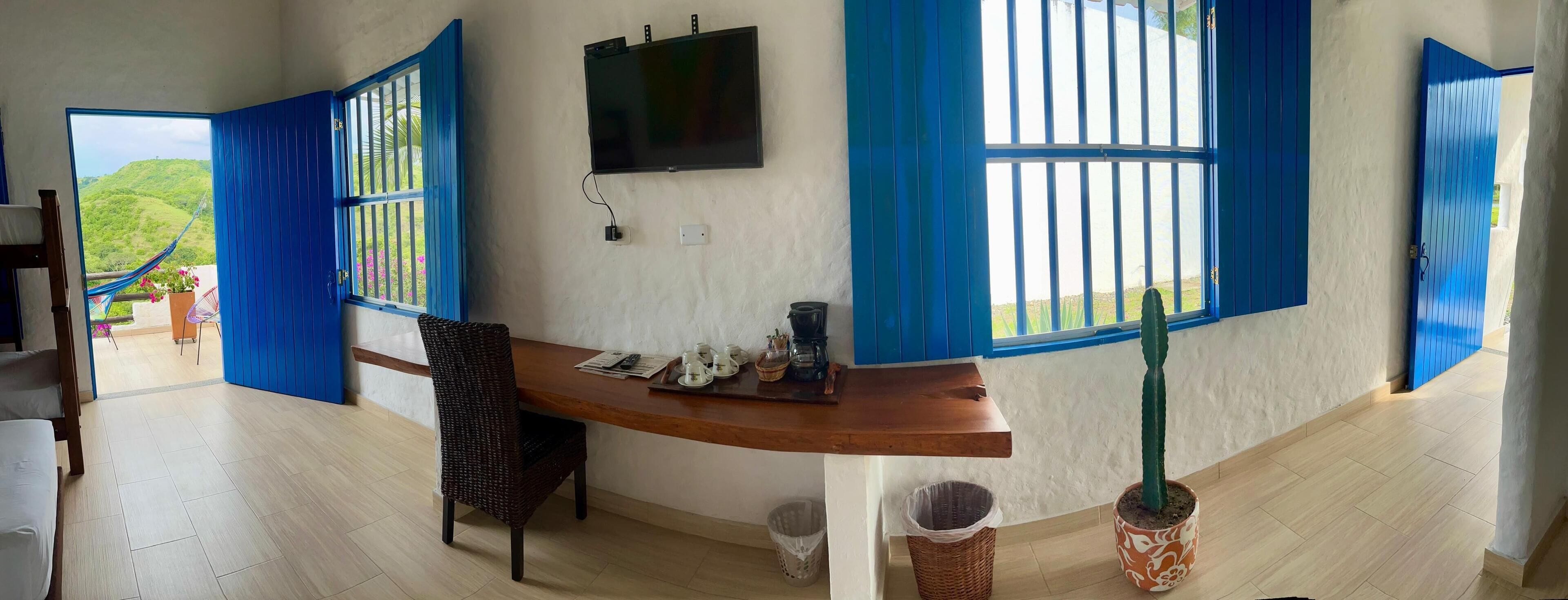 Una habitación con un escritorio de madera, una silla, un televisor y una ventana con contraventanas azules.