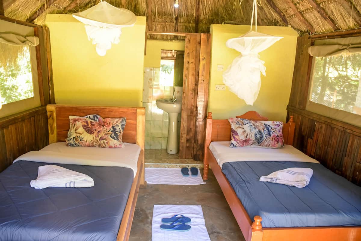 A room with two beds, each with a pillow and a folded towel. There is a window on the left side, and a door leading to a bathroom.