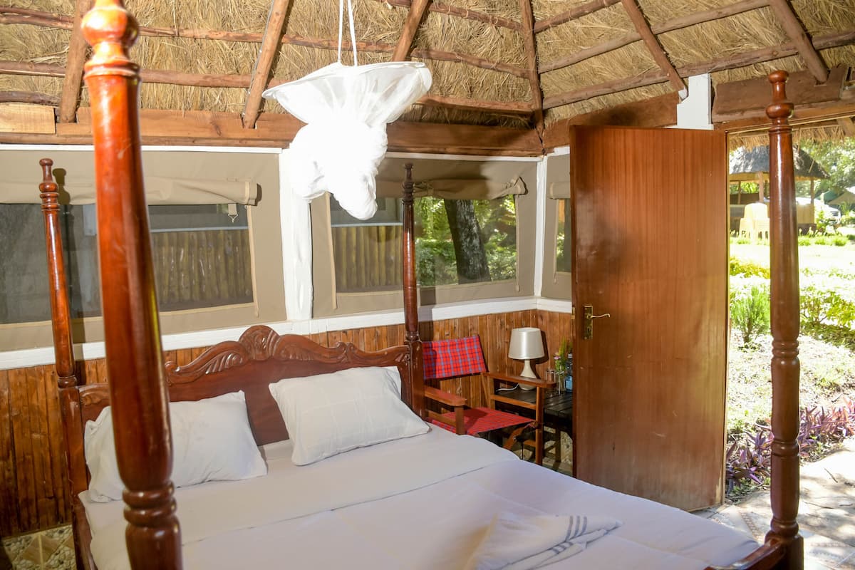 A four-poster bed with a canopy, a wooden nightstand, a chair with a red and black checkered cushion, and a window with a view of greenery.