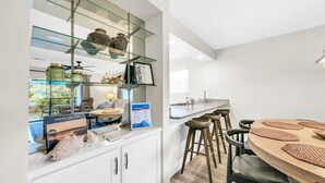 A modern kitchen with a glass shelf displaying decorative items, a wooden dining table with stools, and a view of the outdoors. - 🐟Bikes Included-Cassine Gardens 273-30A (Santa Rosa Beach)