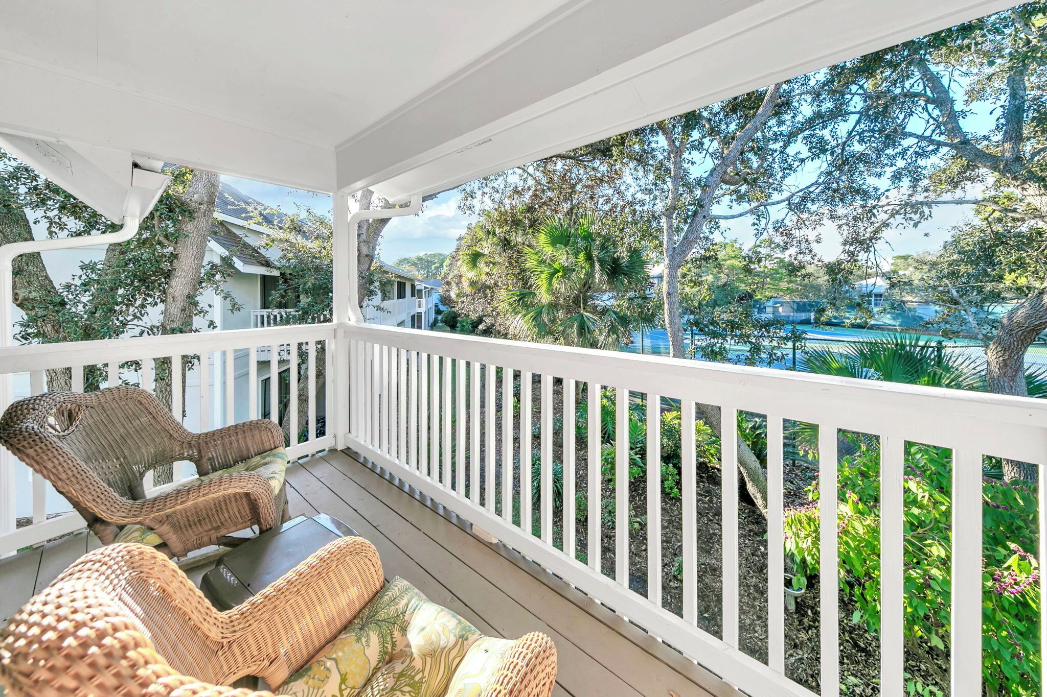 A covered porch with wicker furniture and a white railing, overlooking a residential area with trees and houses.