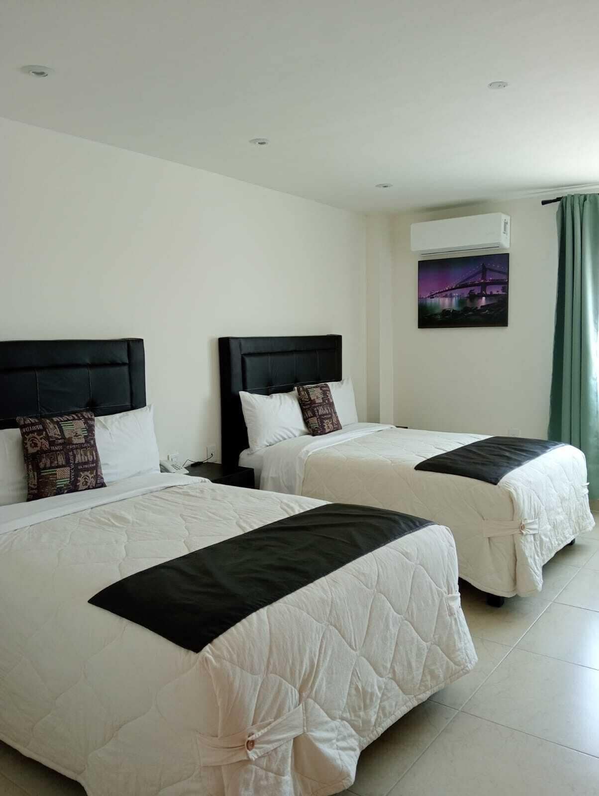 Habitación de hotel con dos camas, cada una con cabecero negro y ropa de cama blanca. Se observa un aire acondicionado de pared y un cuadro con una imagen de un puente.