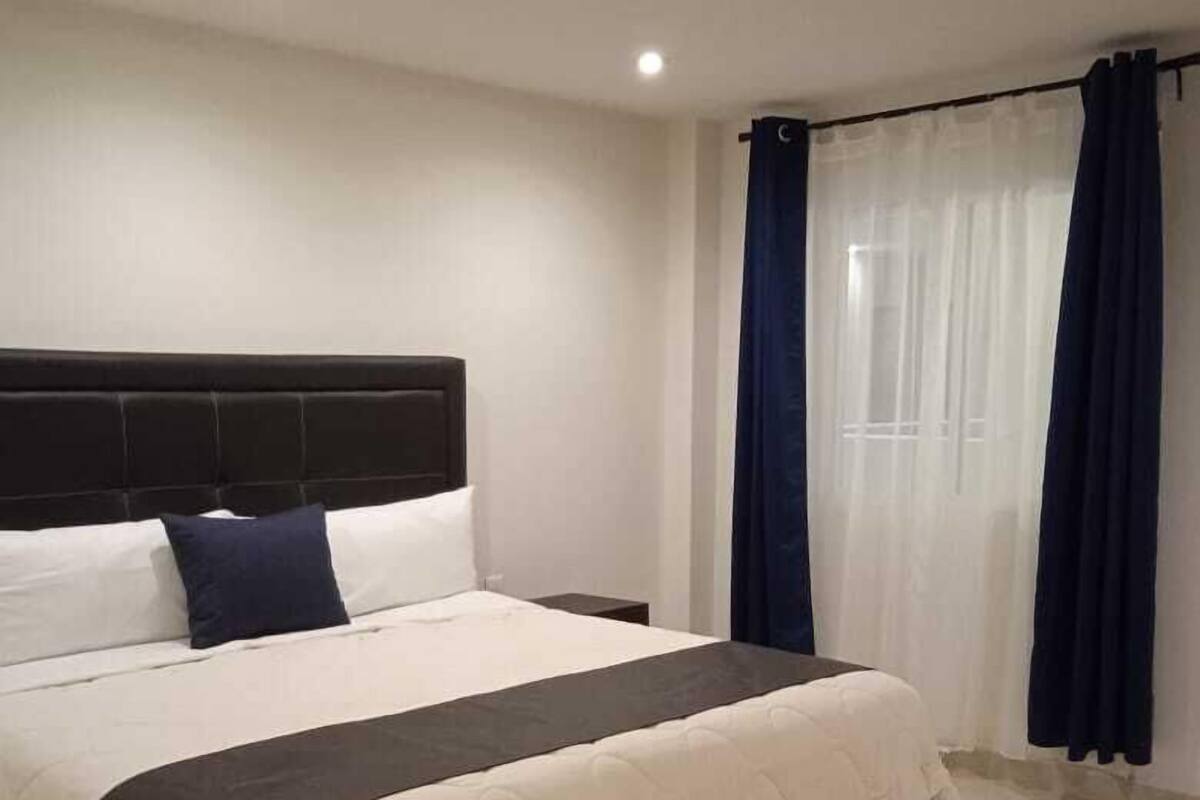 Ein ordentlich bezogenes Bett mit dunklem Kopfteil, weißer Bettwäsche und einer dunklen Tagesdecke.