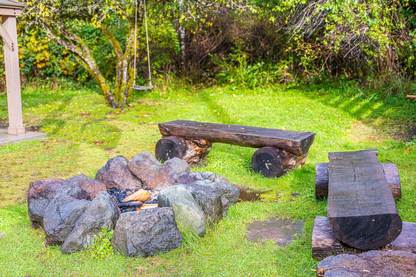 A rustic outdoor seating area with a fire pit surrounded by rocks and logs, set in a lush green garden.
