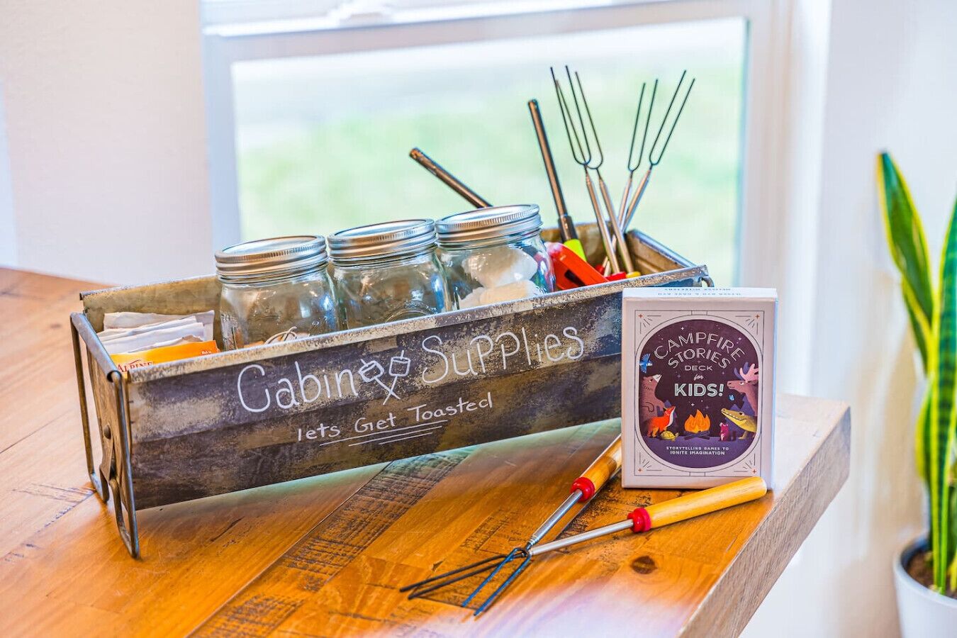 Una caja de madera con la etiqueta 'Suministros para la cabaña', con frascos y utensilios de cocina, y un libro 'Mazo de relatos junto a la fogata' sobre una mesa.