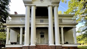 A large, stately building with a central entrance flanked by columns, a covered porch, and a brick pathway leading to the door. - Historic suite 3 (Magnolia)