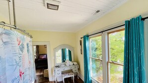 A room with a wooden floor, a white dresser with a mirror, a window with teal curtains, and a shower curtain with a tropical print. - Historic suite 3 (Magnolia)