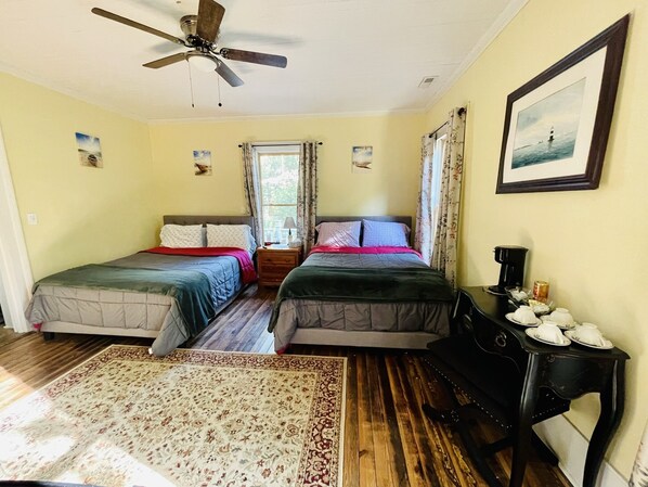 A room with two beds, a ceiling fan, a window with curtains, a framed picture on the wall, and a small table with cups and a coffee pot.