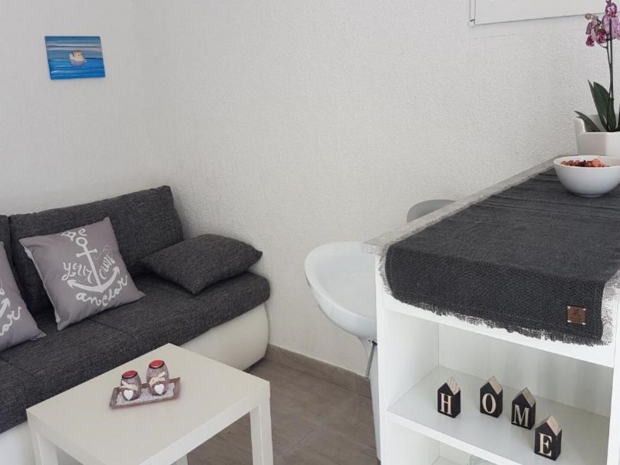 Un salon moderne avec un canapé gris, une petite table blanche agrémentée d’objets décoratifs, des toilettes blanches et une étagère sur laquelle sont disposés des blocs de construction en forme de maison, formant le mot « MAISON ».