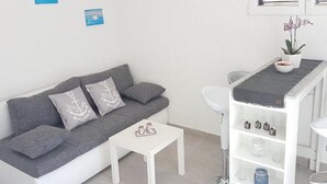 Ein modernes, minimalistisches Zimmer mit einem grauen Sofa, einem kleinen weißen Tisch und einer weißen Regalwand mit Gläsern und einer Pflanze.