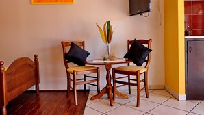 A room with a wooden bed, two wooden chairs, a small round table with a vase, a television on the wall, and a kitchen area with red tiles.