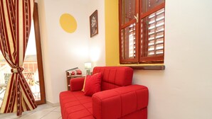 Un divano rosso in una stanza con una finestra, una lampada e un quadro incorniciato al muro.