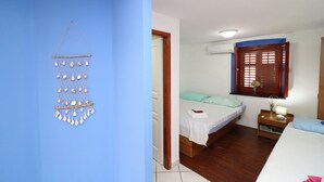 Ein Zimmer mit blauer Wand, einem Bett mit grünem Kopfkissen, einem Nachttisch mit Lampe und einem Fenster mit Holzjalousien.