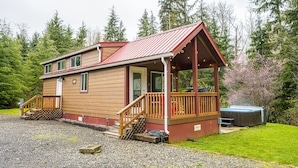 A wooden cabin with a red roof, a porch, and a hot tub in a forested area.