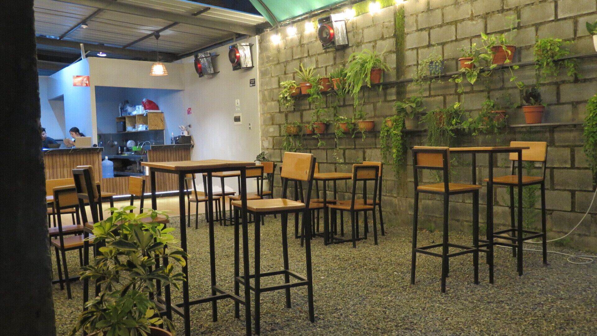 A café with wooden tables and chairs, a brick wall with potted plants, and a visible kitchen area.