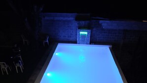 A rectangular swimming pool with a blue illuminated edge, surrounded by a brick wall and a grassy area with chairs.
