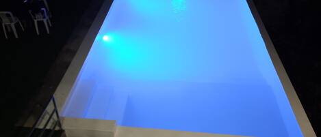 A rectangular swimming pool with a blue illuminated edge, surrounded by a brick wall and a grassy area with chairs.